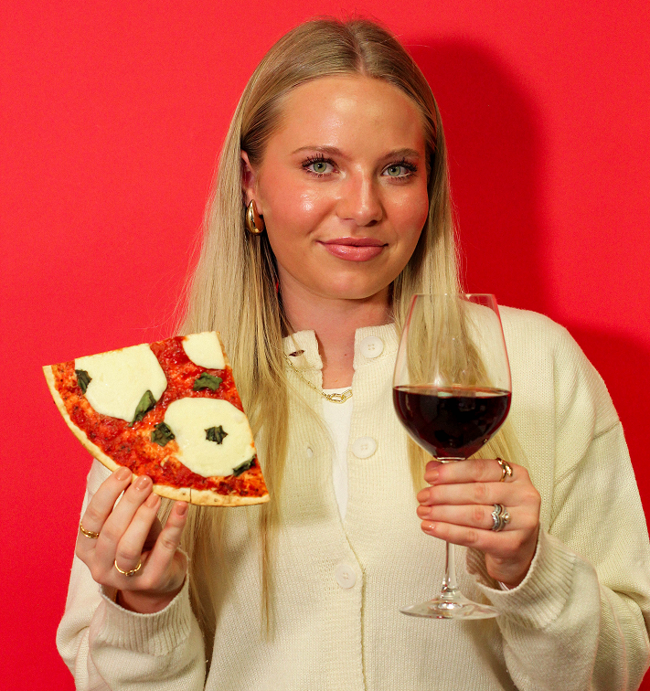 Woman holding a slice of pizza and a glass of red wine against a red background